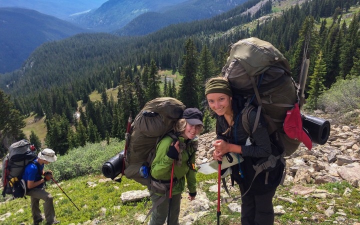 three people with backpacks make their way up a rocky and grassy incline